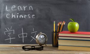 Student learning Chinese with books, pencils, clock, reading glasses and an apple in front of chalkboard with Mandarin text.